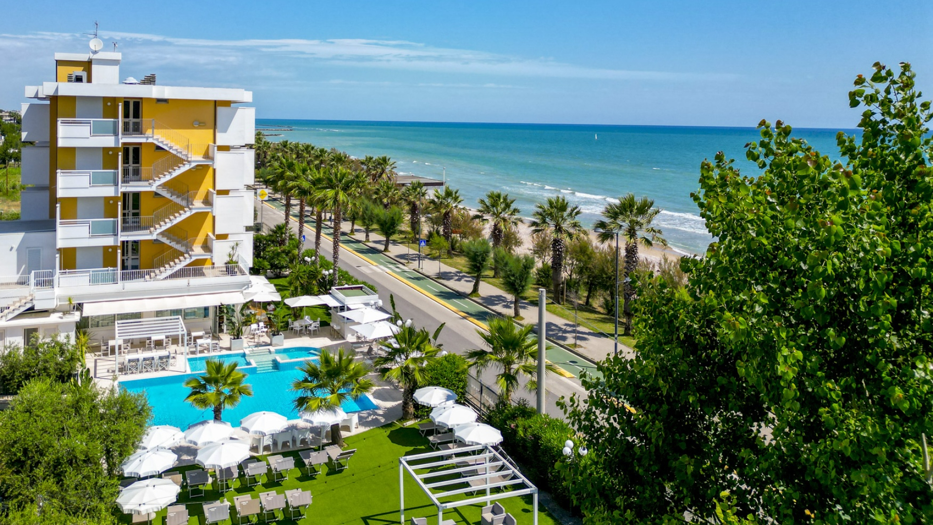 Hotel con piscina vicino al mare, circondato da palme e ombrelloni bianchi.