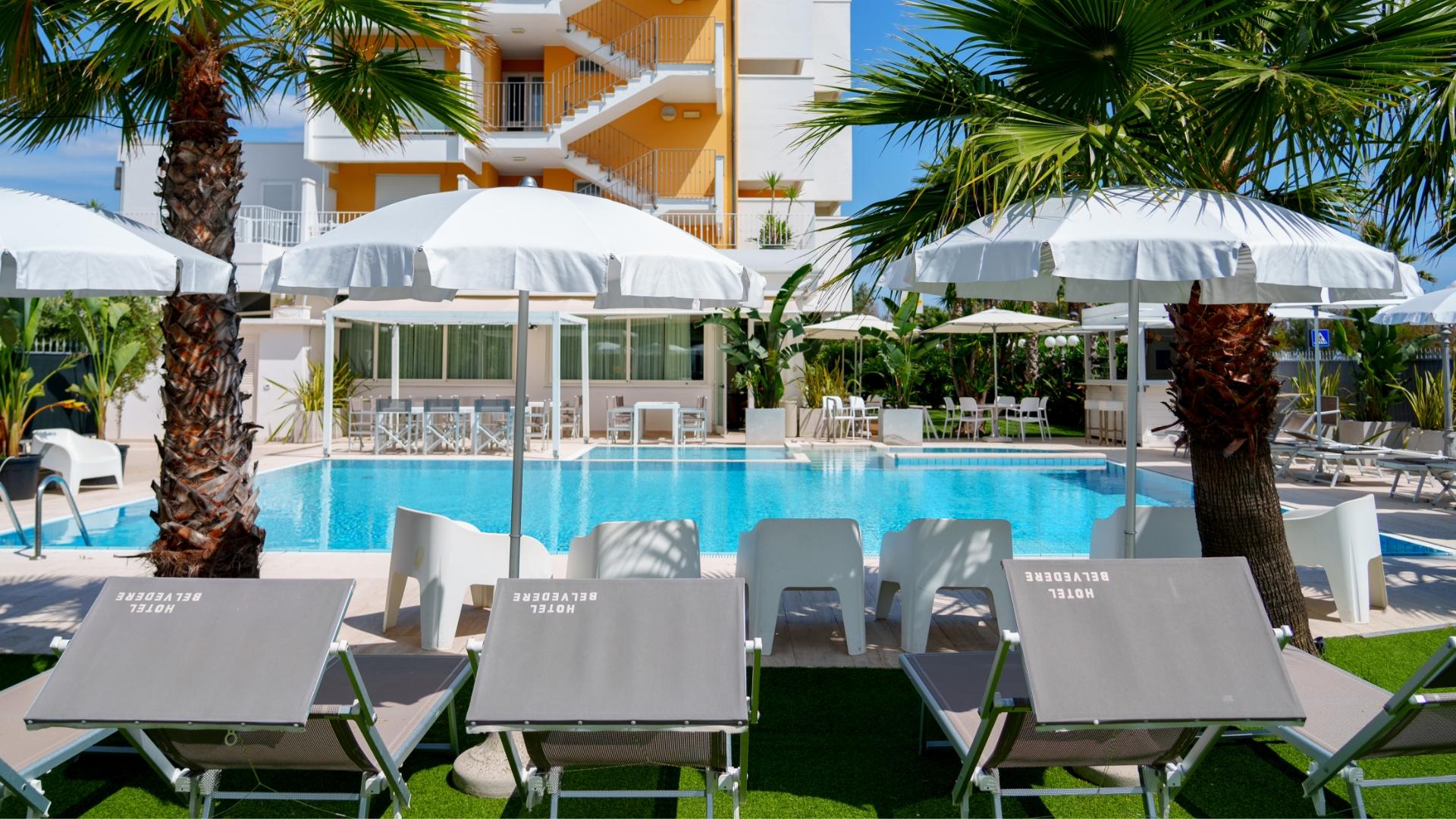 Piscina all'aperto con lettini e ombrelloni bianchi, circondata da palme.