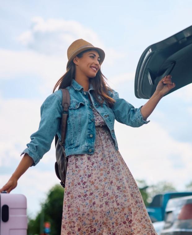 Donna con cappello e valigia apre il bagagliaio dell'auto, sorridente.