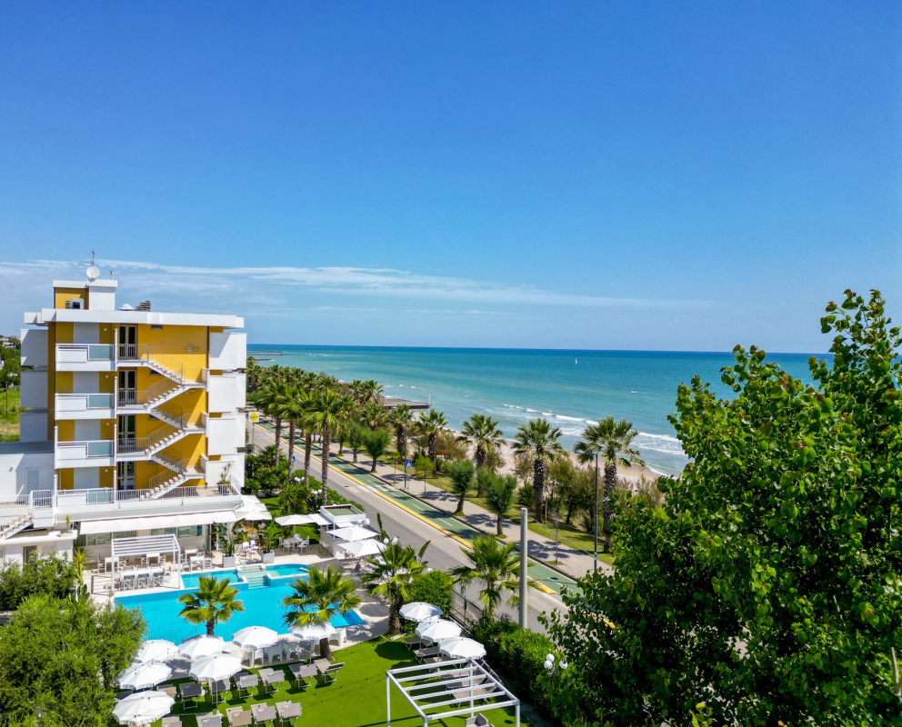 Hotel con piscina vicino al mare, circondato da palme e ombrelloni bianchi.